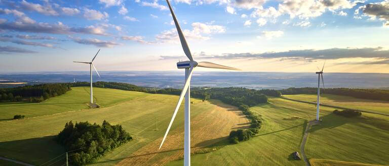 In einer weitläufigen, grünen Landschaft stehen mehrere große Windräder unter einem teils bewölkten Himmel.