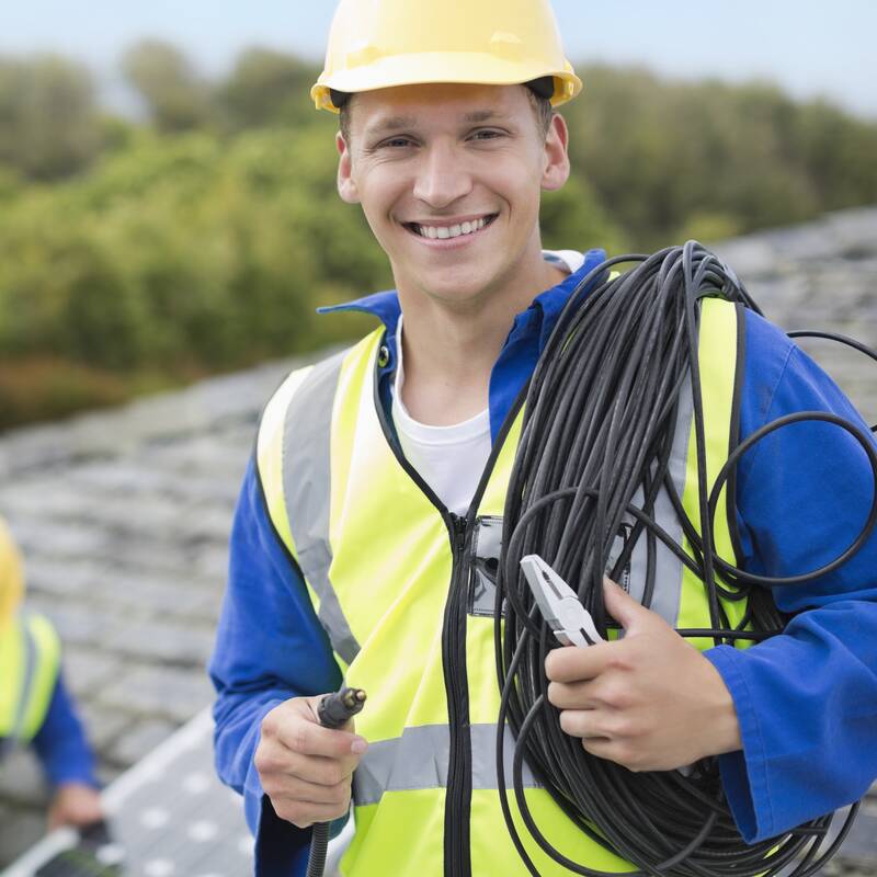 Zwei Elektriker mit Helm auf einem Dach bei der Installation einer Solarstromanlage. Der vordere Mann trägt Kabel