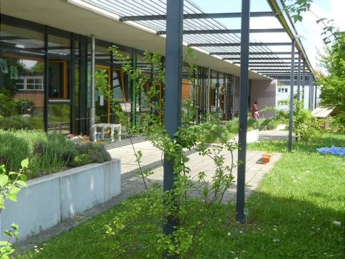 Ein moderner Gebäudebereich mit großen Fenstern und einem überdachten Gehweg ist von grünen Pflanzen und Rasenflächen umgeben.