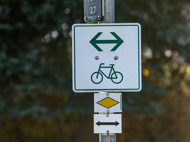 Ein weißes Schild mit einem grünen Fahrrad und Richtungspfeil in beider Richtungen als Wegweiser für einen Radweg. Das Schild ist an einem Laternenpfosten befestigt.